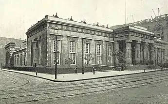 An Egyptian-Revival-style building on a street corner; there are doric columns on the façade to the right of the image. There are men on the street, and work is being done using cranes, on an adjacent building out of frame to the right.