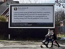 A billboard shows a paragraph of text by Boris Johnson, presented as a tweet.  A woman and a girl, pushing a baby-buggy walk past, looking straight ahead.