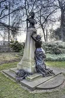 A bronze and stone memorial to Arthur Sullivan. A bronze bust of Sullivan stands on a granite pedestal. A figure of a crying muse leans against the plinth. On the base, bronze sculptures of sheet music, the masks of comedy and tragedy and a mandolin.