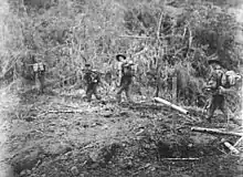 Soldiers patrol through a jungle setting, passing a makeshift grave marker made out of bamboo