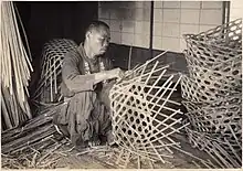 A black and white photograph of a bald-headed Japanese man, crouched on the floor weaving a large basket.
