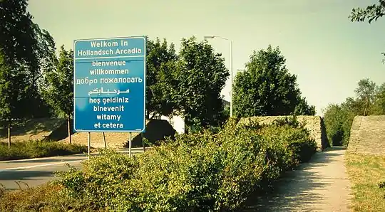 "Welcome" highway sign in several languages, next to a footpath through a wall of sandbags