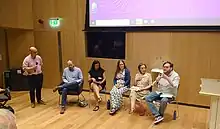 Five people seated, one speaking into a microphone. A sixth person is stood to the side holding another microphone.