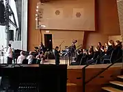 At right a woman in black stands in front of many children dressed in black, sitting in three rows of chairs on a large stage. In the background musicians play a grand piano, drums and guitars. At left a woman stands at a microphone, with several adults seated nearby. The stage opens to the outside at far left.