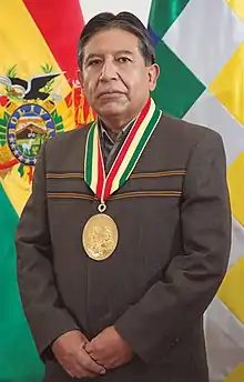 Full body portrait of David Choquehuanca, flanked by the Bolivian flag on the left and the Wiphala on the right.