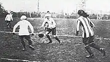 Black and white photo with three players in a white kit, and one player in a striped kit, with a football on a muddy pitch.