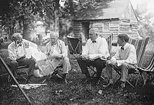 Each of the men sits in a chair outdoors in the grass. Edison reads from a newspaper while the others look at him.