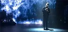 Nottet singing into a microphone against a blue-green background, wearing all-black clothing.