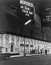 Night. Plain-looking theatre front from across the street, at an angle. Under the marquee, which is as wide as the theatre, a dozen or so people standing in front of the entrance are in bright light. Atop the building, a large sign announces the theatre's name and the play's name (abbreviated as Oh Dad Poor Dad, etc.). Everything else above the marquee, including nearby and distant buildings, is in relative darkness, but the street lights and illuminated signs and windows make it visible.