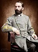 Colorized photo of a bearded man seated in a chair. He wears a gray military uniform with two rows of gold buttons and yellow frogging on the sleeves.