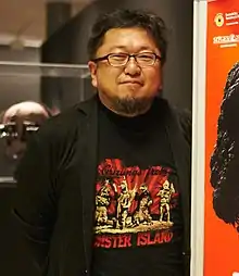 Higuchi stands next to a poster of his 2016 film Shin Godzilla. He wears a T-Shirt emblazoned with the text "Greetings from Monster Island", featuring an illustration of Megalon, Gigan, Godzilla, and Jet Jaguar posing in the 1973 film Godzilla vs. Megalon.