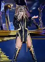 Taylor Swift singing on a microphone, dressed in a black suit with golden accents