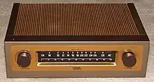 A small rectangular brown box with a metallic coppery top on a beige carpet. On its front are two dials and a tuner with a range between 88 and 108